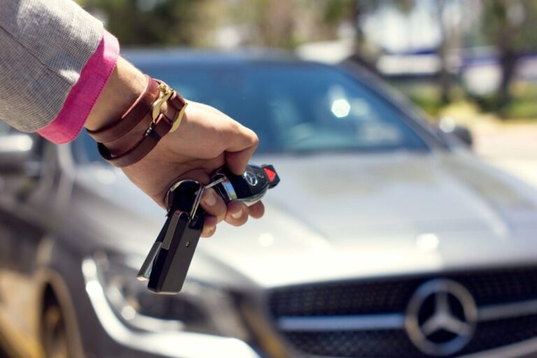 A person holds a Mercedes-Benz car key fob in front of a silver Mercedes-Benz vehicle, with the car logo visible on the grille.