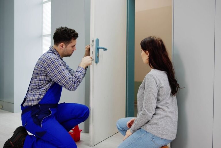 A man in blue overalls repairs a door lock with a screwdriver while a woman sits nearby watching.