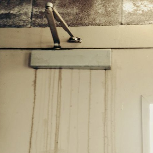 A metal door closer mounted at the top of a beige door, with several water stains running downward from the device.