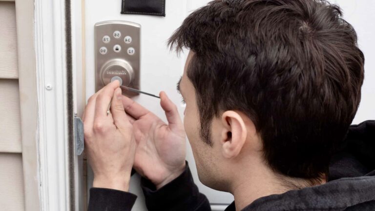 A person is picking the lock of a door using a thin tool, focusing on a keypad lock system mounted next to a white door frame.