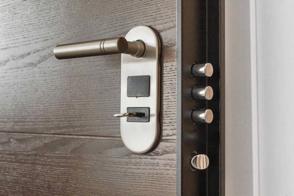 A metal door handle and lock with a key inserted, shown on a dark wooden door that is slightly open, revealing security bolts on the door frame.