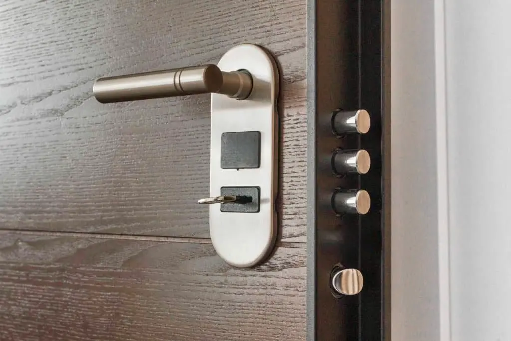 A metal door lock with a key inserted, a door handle, and three locking bolts visible on the edge of a wooden door.
