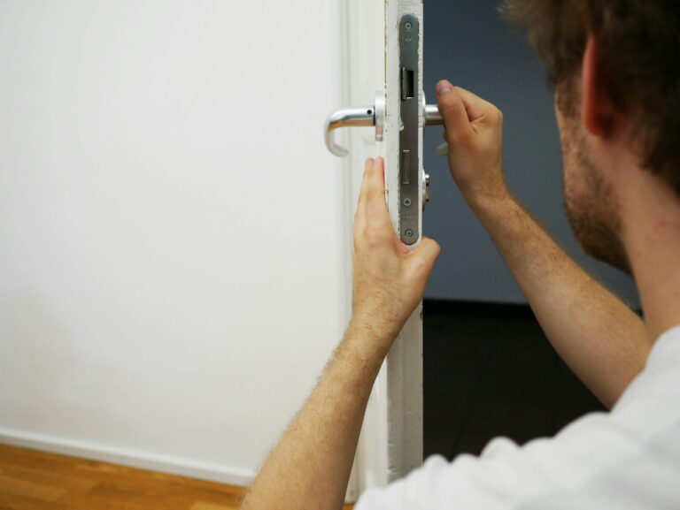 A 24-hour emergency locksmith in Vancouver is installing or repairing a metal door lock using a screwdriver on a white door, with part of their face and upper body visible, and a blank wall in the background.