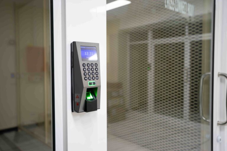 A biometric fingerprint scanner with a keypad and high-security locks is mounted on a wall next to a glass door in an office setting. The device display shows the time as 12:31, demonstrating the kind of advanced security solutions provided by a commercial locksmith in Vancouver.