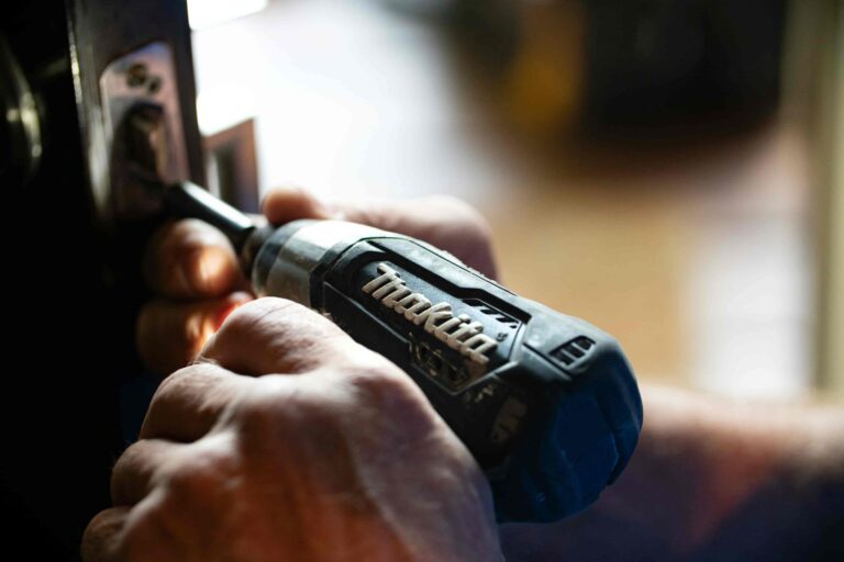 Close-up of hands using a Makita power drill to install or remove a screw from a metal door latch, demonstrating professional locksmith services in Vancouver.